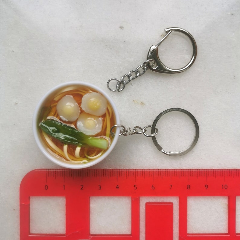 May include: A white bowl of ramen noodles with three egg dumplings and a green onion garnish. The bowl is attached to a silver key chain.