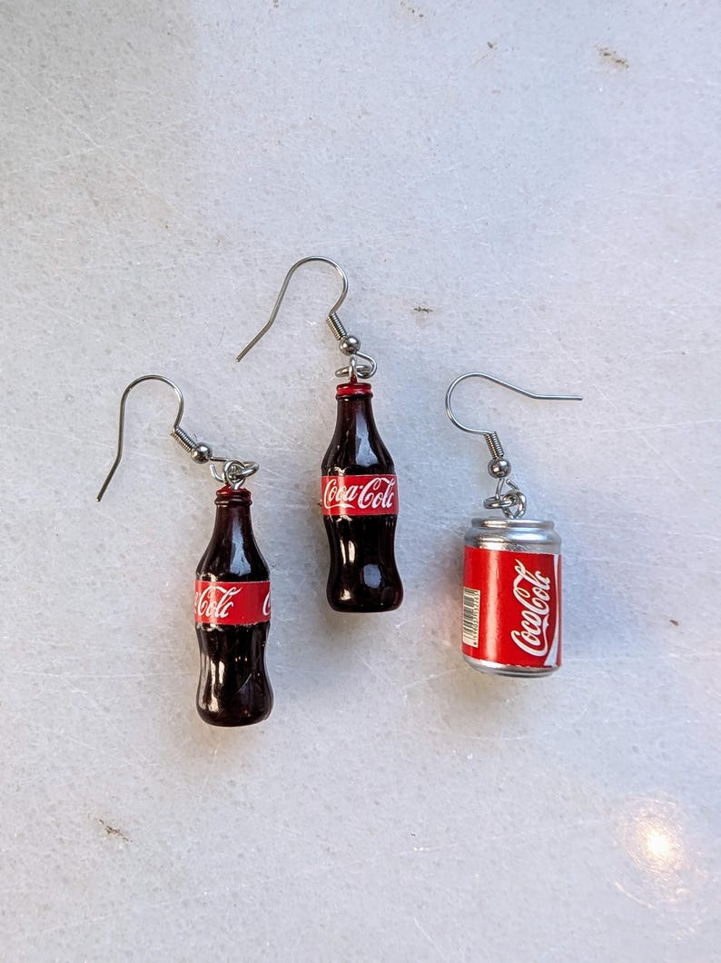 Puede incluir: Un par de pendientes de plata con botellas de Coca-Cola en miniatura y una lata. Las botellas son marrones con etiquetas rojas y blancas que dicen "Coca-Cola". La lata es plateada con una etiqueta roja y blanca que dice "Coca-Cola".