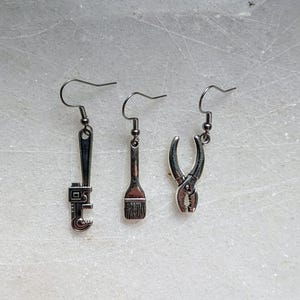May include: Three silver-toned earrings, each with a different tool charm: a wrench, a paintbrush, and pliers. The earrings hang from simple ear wires. The tools are detailed and appear to be miniature replicas.