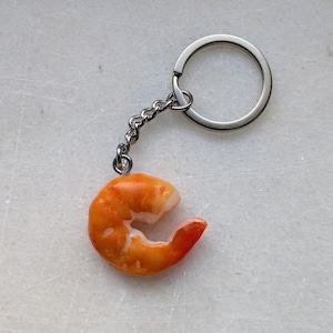 May include: A novelty keyring featuring a realistic, orange and white shrimp charm. The shrimp is attached to a silver-coloured chain and keyring. A fun and unique accessory.