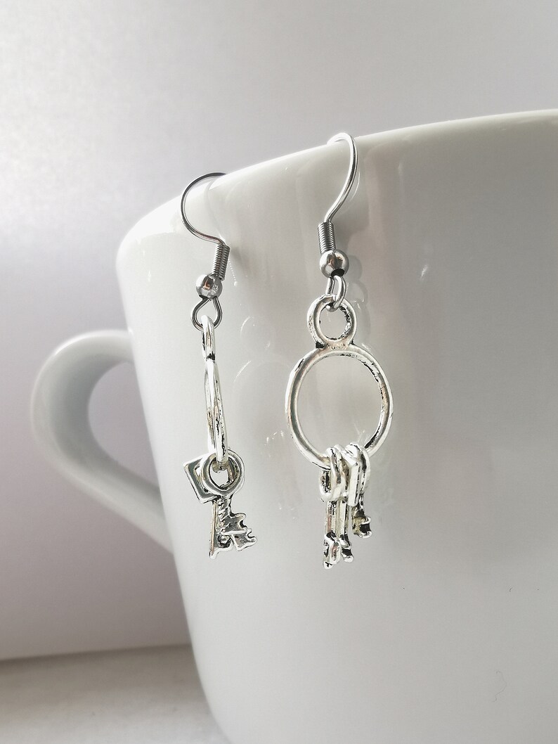 May include: A pair of silver key-shaped earrings. One earring features a single key charm, while the other has a cluster of three keys hanging from a hoop.