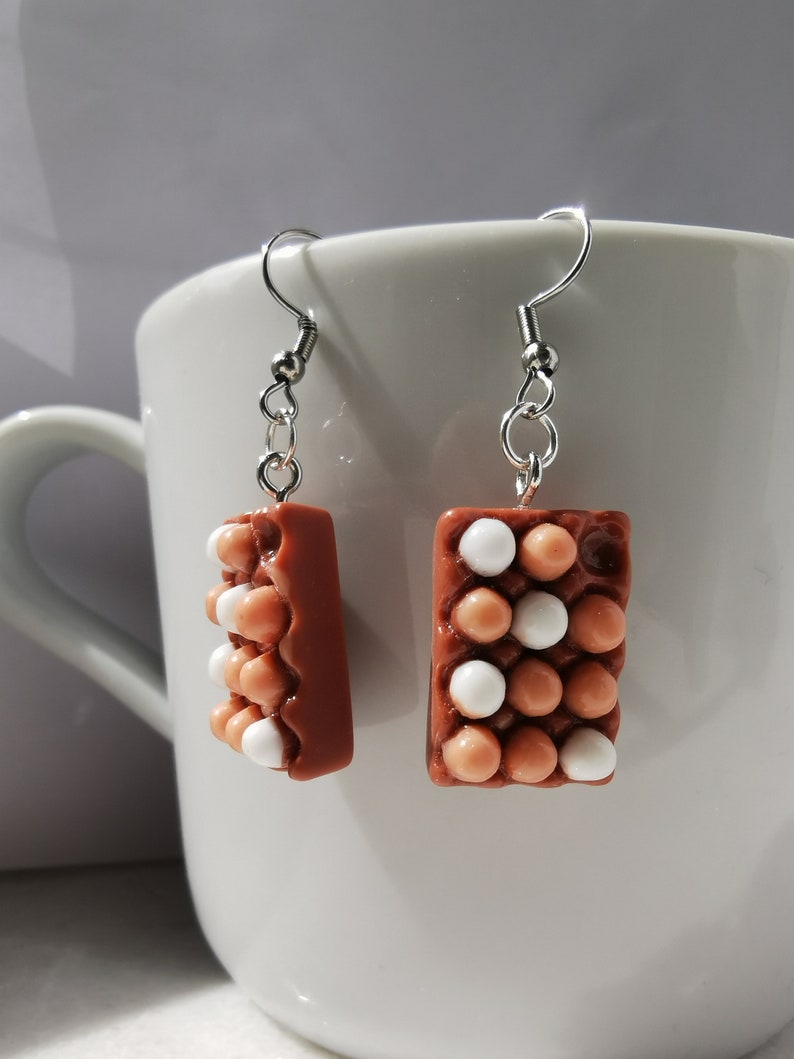 Peut inclure: Une paire de boucles d'oreilles pendantes avec un motif de bo&icirc;te &agrave; &oelig;ufs marron avec des &oelig;ufs blancs et marrons. Les boucles d'oreilles sont en p&acirc;te polym&egrave;re et ont des crochets argent&eacute;s.