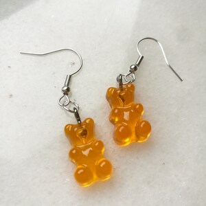 K&ouml;nnte beinhalten: Ein Paar Ohrringe mit H&auml;ngern in Form von Gummib&auml;rchen in leuchtend orange. Die Ohrringe sind aus silberfarbenem Metall.