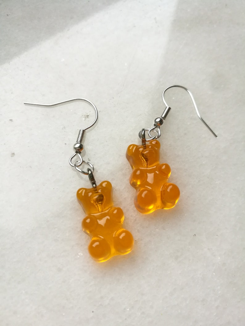 K&ouml;nnte beinhalten: Ein Paar Ohrringe mit H&auml;ngern in Form von Gummib&auml;rchen in leuchtend orange. Die Ohrringe sind aus silberfarbenem Metall.