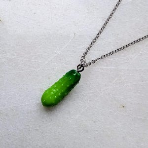 May include: A silver chain necklace with a bright green pickle pendant. The pickle has a textured surface and is suspended from a small loop attached to the chain. The necklace is displayed against a white background.