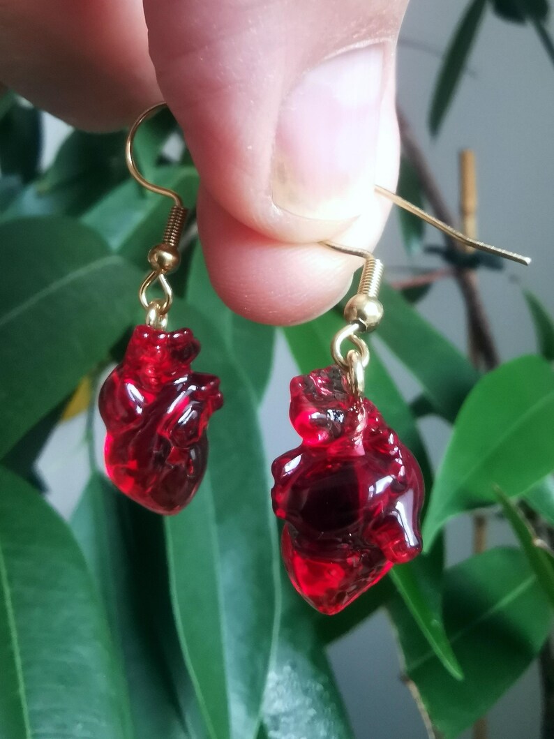 May include: A pair of gold heart-shaped earrings with red glass hearts. The hearts are detailed and realistic.