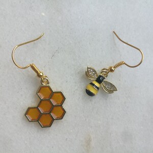 K&ouml;nnte beinhalten: Ein Paar goldene Ohrringe mit einem Wabenmuster und einem Bienenmuster. Der Wabenohrring ist orange und der Bienenohrring ist gelb mit schwarzen Streifen.