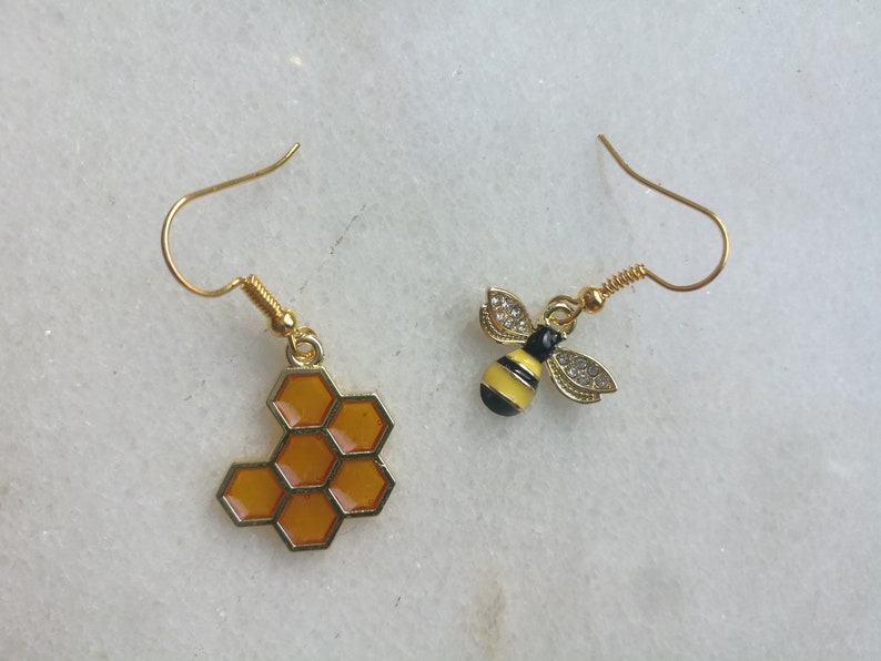 K&ouml;nnte beinhalten: Ein Paar goldene Ohrringe mit einem Wabenmuster und einem Bienenmuster. Der Wabenohrring ist orange und der Bienenohrring ist gelb mit schwarzen Streifen.
