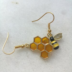 K&ouml;nnte beinhalten: Goldene Bienen- und Wabenohrringe. Die Biene hat schwarze und gelbe Streifen und klare Strasssteine. Die Wabe ist hellbernsteinfarben.