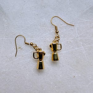 May include: A pair of gold-colored earrings featuring miniature coffee maker charms. The earrings have a hook design for pierced ears. The coffee maker charms are detailed with handles and a classic espresso pot shape. The earrings are displayed on a white marble surface.