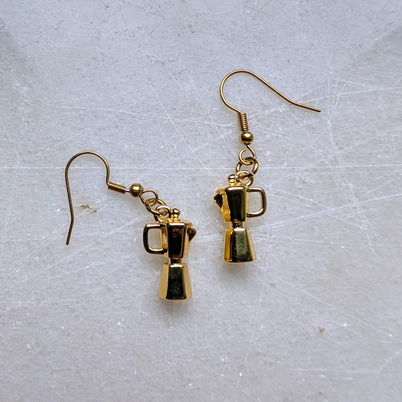 May include: A pair of gold-colored earrings featuring miniature coffee maker charms. The earrings have a hook design for pierced ears. The coffee maker charms are detailed with handles and a classic espresso pot shape. The earrings are displayed on a white marble surface.