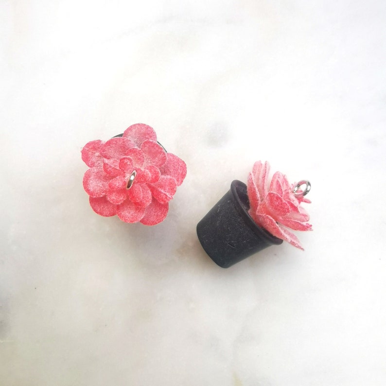 May include: Two small, handmade succulent charms. One is a red flower shape, the other is in a black pot. Both have a silver-colored loop for attaching to jewelry or other crafts. The succulents are on a white surface.