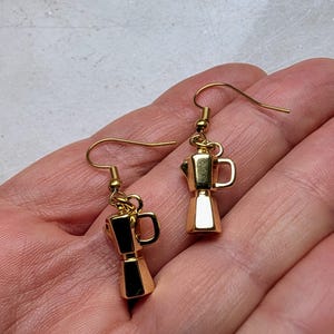 May include: A pair of gold-colored earrings featuring miniature moka pot charms. The earrings have a classic coffee maker design with a handle and spout. The charms are suspended from gold-colored ear wires, creating a stylish accessory for coffee lovers.