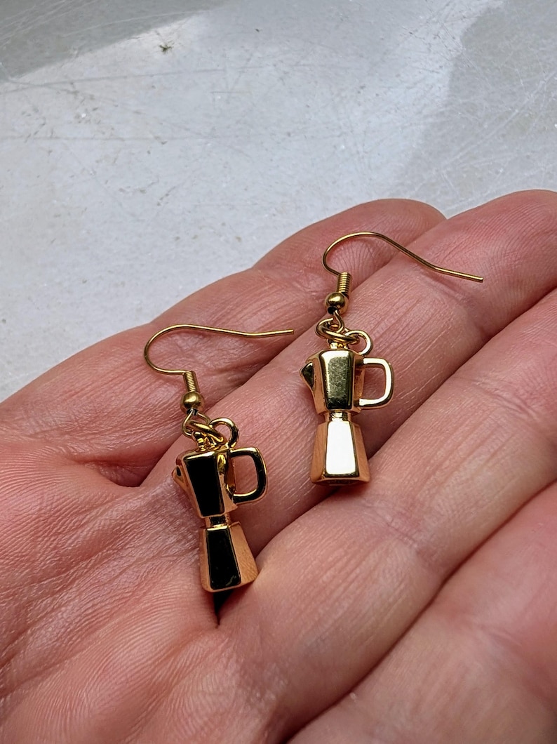 May include: A pair of gold-colored earrings featuring miniature moka pot charms. The earrings have a classic coffee maker design with a handle and spout. The charms are suspended from gold-colored ear wires, creating a stylish accessory for coffee lovers.