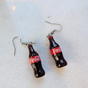 Puede incluir: Un par de pendientes de aro de plata con botellas de Coca-Cola en miniatura con etiquetas rojas y letras blancas.