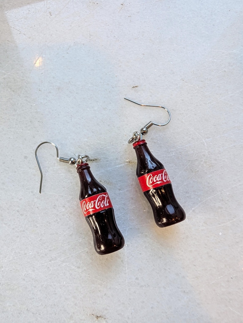 Puede incluir: Un par de pendientes de aro de plata con botellas de Coca-Cola en miniatura con etiquetas rojas y letras blancas.