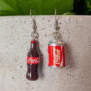 Puede incluir: Un par de pendientes de plata con encantos en miniatura de botella y lata de Coca-Cola. La botella es marr&oacute;n con una etiqueta de Coca-Cola roja. La lata es plateada con una etiqueta de Coca-Cola roja.