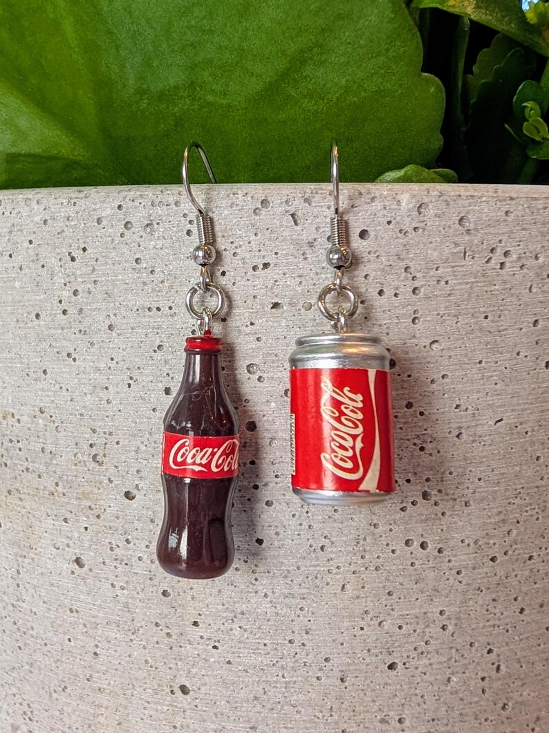 Puede incluir: Un par de pendientes de plata con encantos en miniatura de botella y lata de Coca-Cola. La botella es marr&oacute;n con una etiqueta de Coca-Cola roja. La lata es plateada con una etiqueta de Coca-Cola roja.