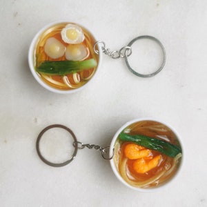 May include: Two white ceramic bowls with a clear liquid and noodles inside, topped with green onions and egg. The bowls are attached to silver keychains.