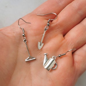 Puede incluir: Tres pendientes de plata con dijes de temática de jardinería. Los pendientes están en una mano, y los dijes son un rastrillo, una pala y una regadera.