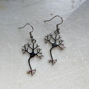 Può includere: Un paio di orecchini a neurone color argento. Ogni orecchino presenta un design dettagliato del neurone con un gancio per orecchie forate. Il design del neurone ha un corpo centrale con dendriti ramificati e un assone.