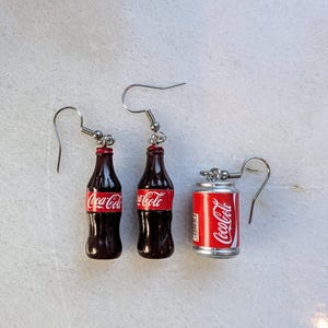Puede incluir: Un par de pendientes de plata con botellas de Coca-Cola en miniatura y una lata. Las botellas son marrones con etiquetas de Coca-Cola rojas. La lata es plateada con una etiqueta de Coca-Cola roja.