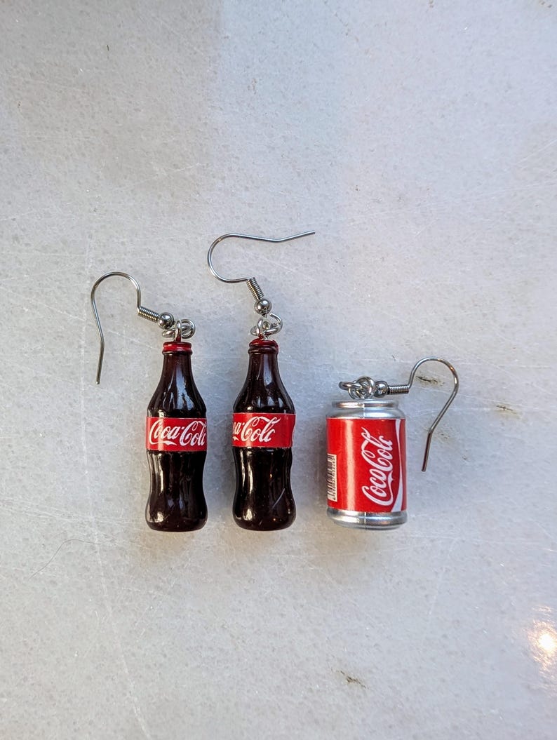 Puede incluir: Un par de pendientes de plata con botellas de Coca-Cola en miniatura y una lata. Las botellas son marrones con etiquetas de Coca-Cola rojas. La lata es plateada con una etiqueta de Coca-Cola roja.