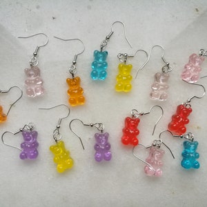 K&ouml;nnte beinhalten: Sortiment an Gummib&auml;rchen-Ohrringen in verschiedenen Farben, darunter Orange, Gelb, Rot, Rosa, Lila, Blau und Klar. Die Ohrringe sind aus klarem Kunststoff und haben silberne Haken.