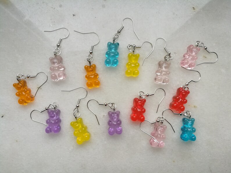 K&ouml;nnte beinhalten: Sortiment an Gummib&auml;rchen-Ohrringen in verschiedenen Farben, darunter Orange, Gelb, Rot, Rosa, Lila, Blau und Klar. Die Ohrringe sind aus klarem Kunststoff und haben silberne Haken.