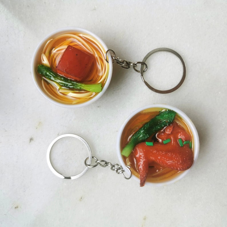 May include: Two keychains featuring miniature bowls of noodles. One bowl has a red meat and green vegetable topping, the other has a brown meat and green vegetable topping.