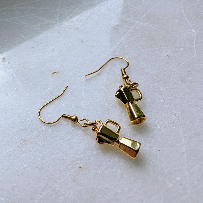 May include: A pair of gold-colored earrings. Each earring features a miniature coffee maker charm with a hook closure. The earrings are displayed on a white surface.