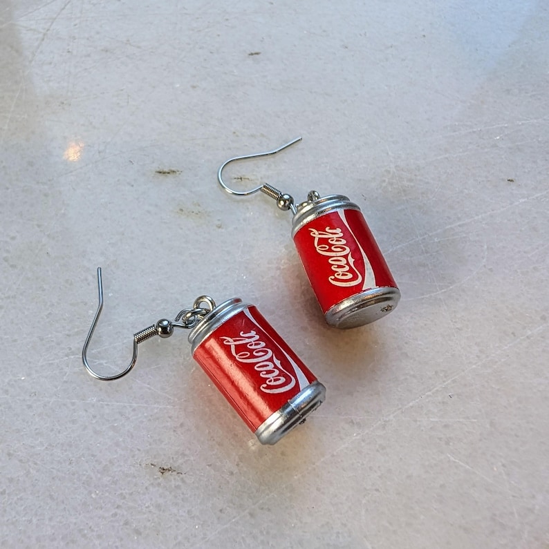 Puede incluir: Un par de pendientes de plata con forma de latas de Coca-Cola. Las latas son rojas con letras blancas que dicen "Coca-Cola".