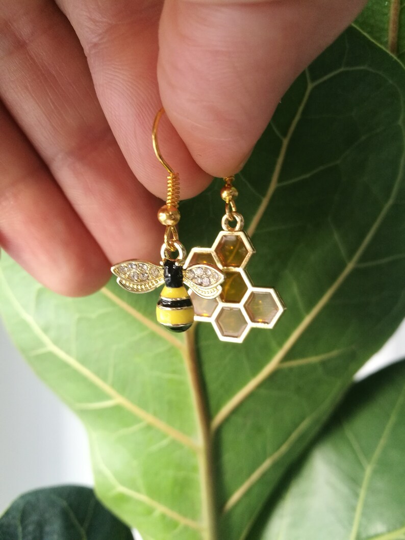 K&ouml;nnte beinhalten: Goldene Bienen- und Wabenohrringe. Die Biene ist schwarz und gelb mit klaren Strasssteinen. Die Wabe ist golden mit klaren Strasssteinen.