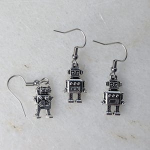 May include: Silver robot earrings with a hook fastening. The robot design has a rectangular body, square head, and small feet. One earring is detached, showing the hook detail. The earrings are displayed on a white marble surface.