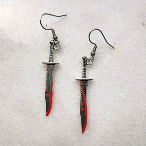 Puede incluir: Un par de pendientes de plata con forma de cuchillos ensangrentados. Los cuchillos tienen un borde dentado y un diseño de salpicaduras de sangre roja.