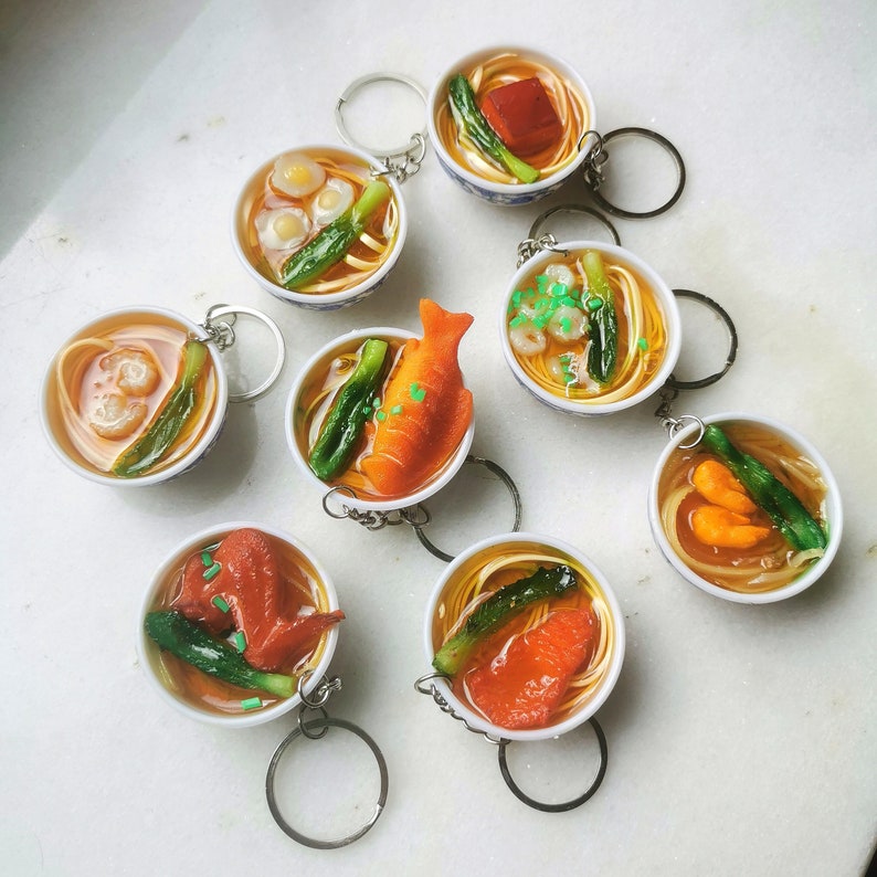 May include: Eight small white bowls with colorful noodle dishes, each with a different topping, including fish, shrimp, and vegetables. The bowls are arranged on a white surface.