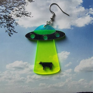 May include: Green, blue, and black UFO earring with a black silhouette of a cow inside. The UFO is hovering over a green field.