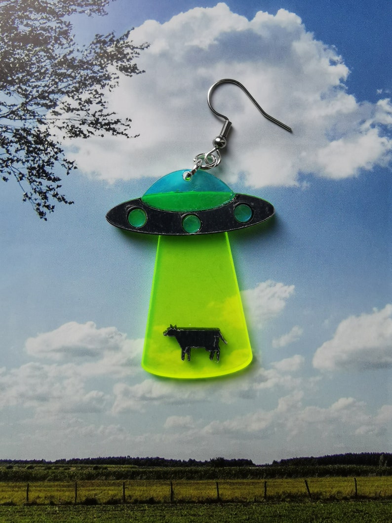 May include: Green, blue, and black UFO earring with a black silhouette of a cow inside. The UFO is hovering over a green field.