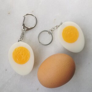 Puede incluir: Tres huevos, un huevo marrón entero y dos huevos blancos cortados por la mitad para mostrar la yema. Las dos mitades de los huevos blancos están unidas a llaveros.