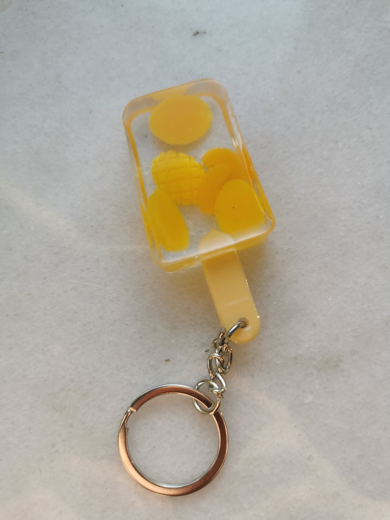 以下が含まれることがあります： 透明なレジン製のトップと白いプラスチック製のハンドルが付いた黄色のアイスキャンディーのキーホルダー。レジンには黄色とオレンジ色の形が入っています。