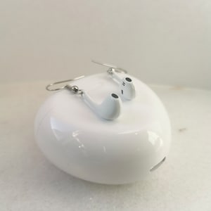 Puede incluir: Pendientes blancos con forma de auriculares con ganchos plateados, colocados sobre una funda de carga blanca y redondeada. Los auriculares tienen detalles negros y la letra "R" en uno. Una divertida interpretación de la tecnología moderna.