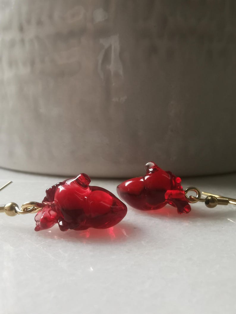 May include: A pair of gold heart-shaped earrings with red glass hearts. The hearts are detailed and have a realistic anatomical look.