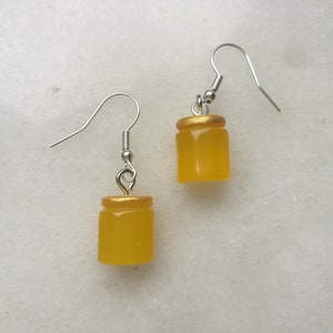 Puede incluir: Un par de pendientes colgantes de color dorado y amarillo con forma de pequeños frascos. Los pendientes tienen ganchos plateados.