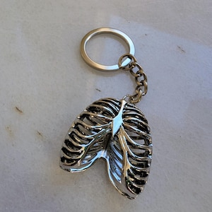 Könnte beinhalten: Silberfarbener Schlüsselanhänger in Form eines Brustkorbs mit einer silbernen Kette und einem Ring.