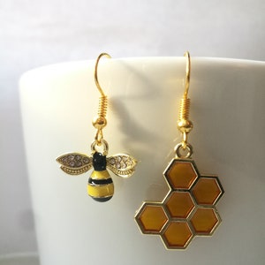 K&ouml;nnte beinhalten: Ein Paar goldene Ohrringe mit einem Bienen- und Wabenmuster. Die Biene ist gelb und schwarz mit klaren Strasssteinen auf den Fl&uuml;geln. Die Wabe ist orange und gold.