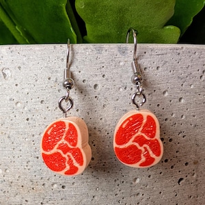 May include: A pair of novelty earrings with a meat design. Each earring has a light pink base with a red, marbled pattern, mimicking a slice of meat. The earrings hang from silver-coloured fish hook ear wires.