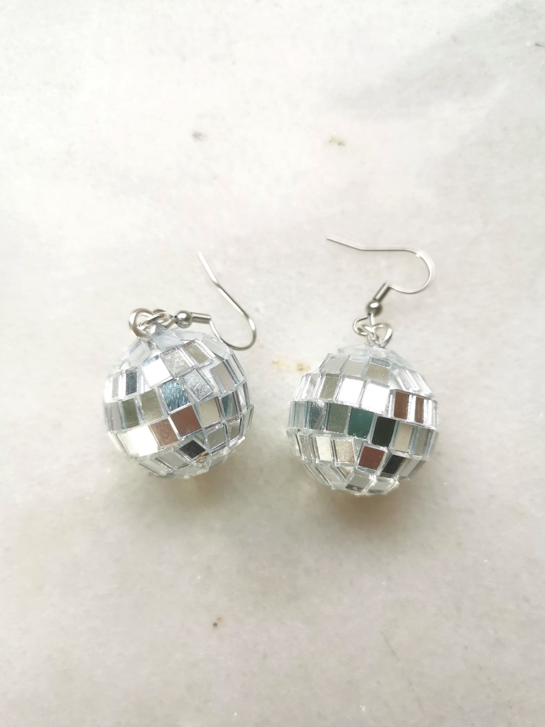 May include: A pair of silver disco ball earrings. The earrings are round and have a mirrored surface. They are hanging from silver hooks.