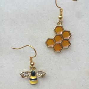 K&ouml;nnte beinhalten: Ein Paar goldene Ohrringe mit einem Bienen- und Wabenmuster. Der Bienenohrring hat einen gelben K&ouml;rper mit schwarzen Streifen und klaren Strasssteinen. Der Wabenohrring ist golden mit einem durchscheinenden orangefarbenen Wabenmuster.