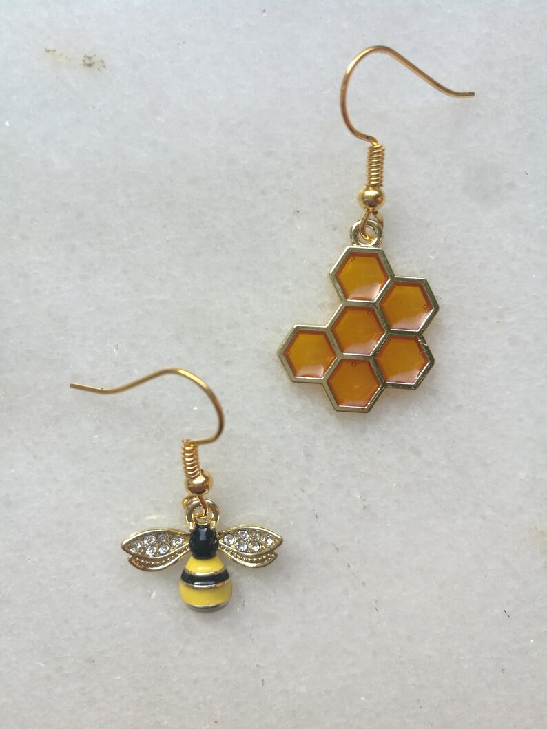 K&ouml;nnte beinhalten: Ein Paar goldene Ohrringe mit einem Bienen- und Wabenmuster. Der Bienenohrring hat einen gelben K&ouml;rper mit schwarzen Streifen und klaren Strasssteinen. Der Wabenohrring ist golden mit einem durchscheinenden orangefarbenen Wabenmuster.