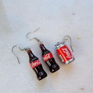 Puede incluir: Un par de pendientes colgantes de plata con botellas de Coca-Cola en miniatura y una lata. Las botellas son de vidrio marr&oacute;n con etiquetas de Coca-Cola rojas y blancas. La lata es de plata con una etiqueta de Coca-Cola roja y blanca.